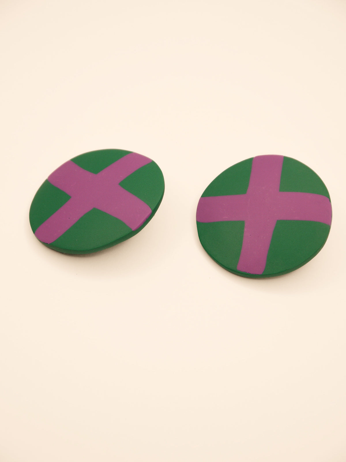 Two round green stud earrings with lilac 'X' design on a beige background