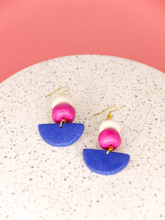 Geometric dangle statement earrings with a blue glitter semi circle, hand rolled beads in pink metallic and white glitter with a brass ball bead hung from gold plated ear wires