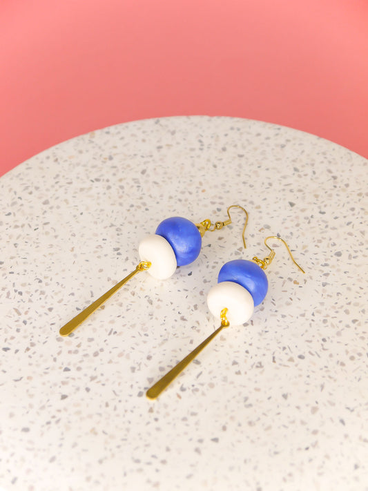 Metallic bead dangle earrings with two hand rolled metallic beads in blue and white glitter with a brass stick charm, hung from gold plated ear wires