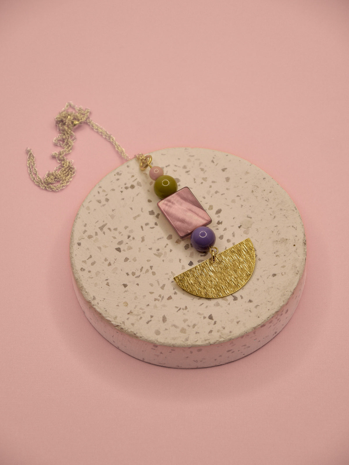 A bohemian-style beaded necklace with a gold chain, featuring a brown mother-of-pearl shell bead, a purple bead, a green bead, and a textured gold half-moon charm, displayed on a round speckled concrete pedestal against a solid pink background.