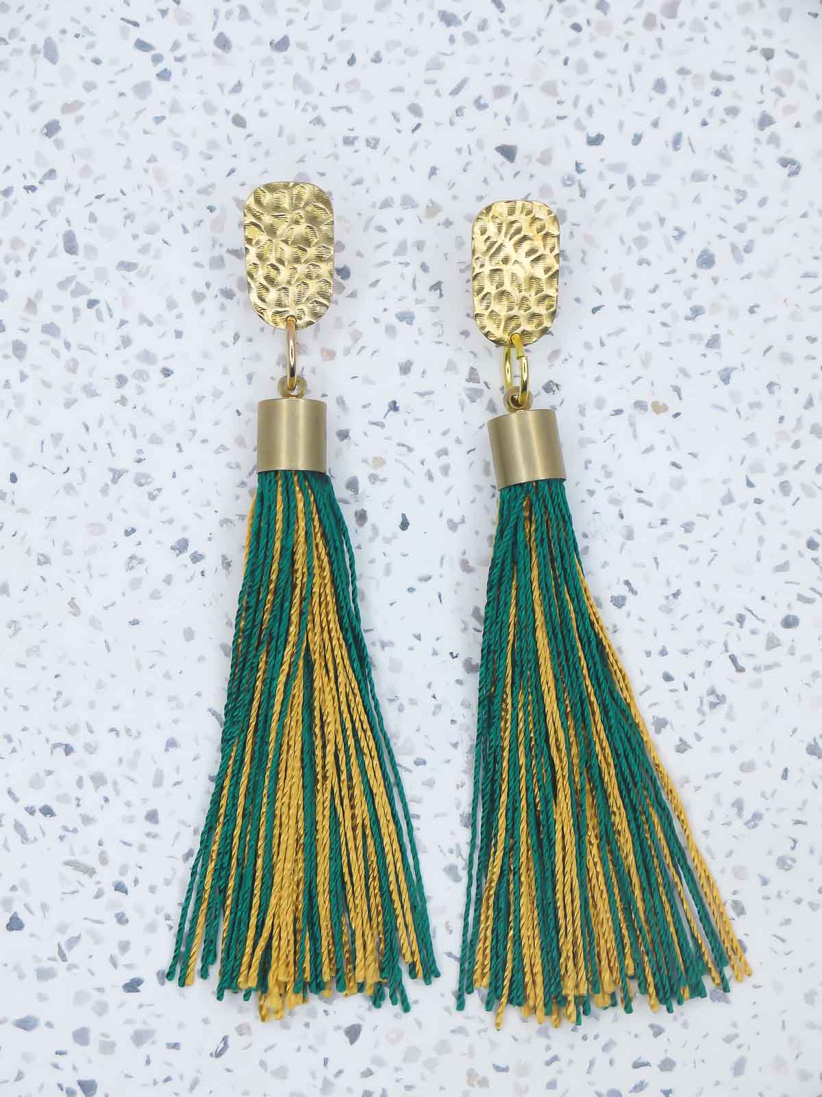 Green and gold tassel earrings with a textured brass stud on a marbled white surface with a pink background