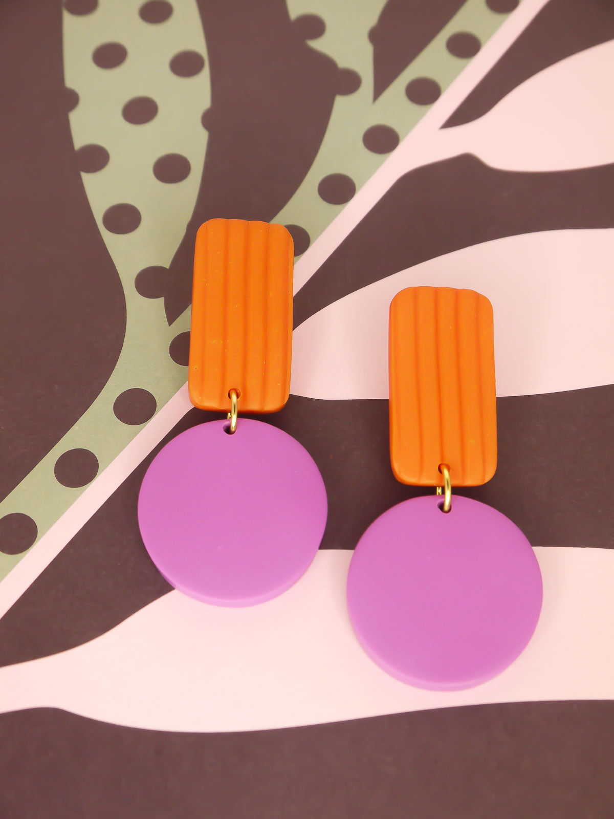 Pair of geometric earrings with orange striped rectangular tops and purple round bottoms on a patterned background.