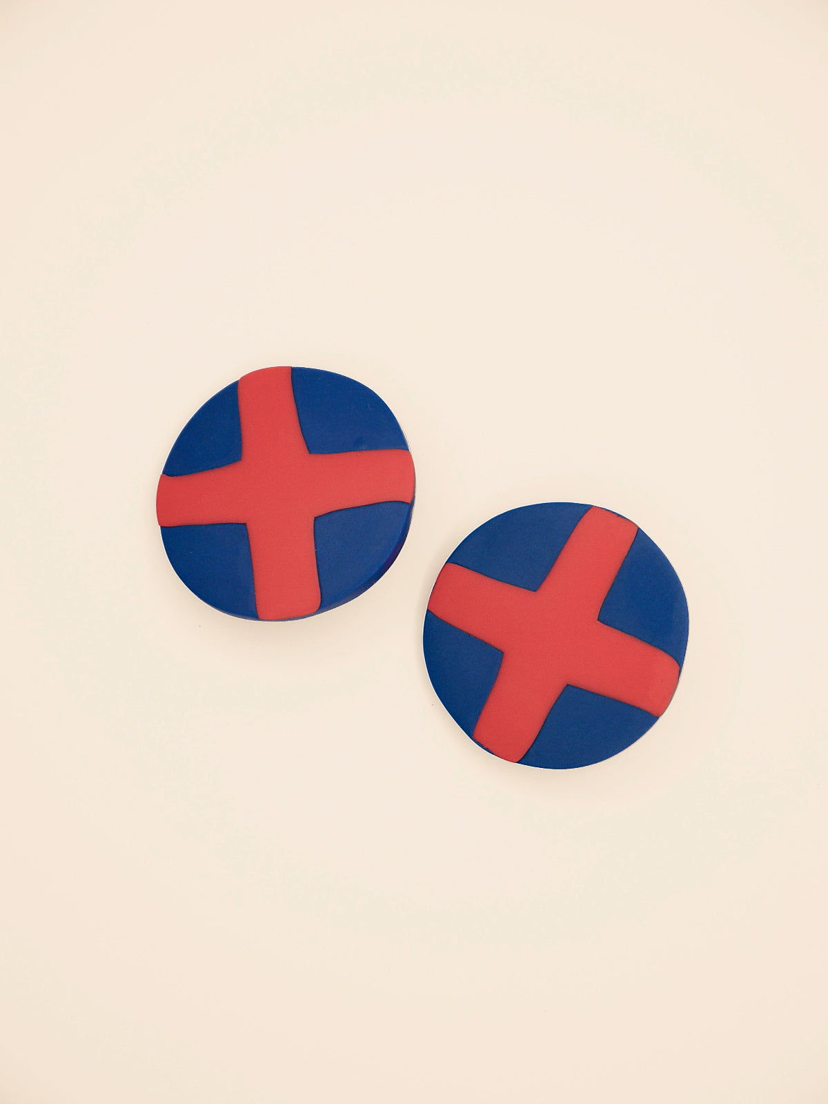 Two round blue stud earrings with red 'X' design on a beige background