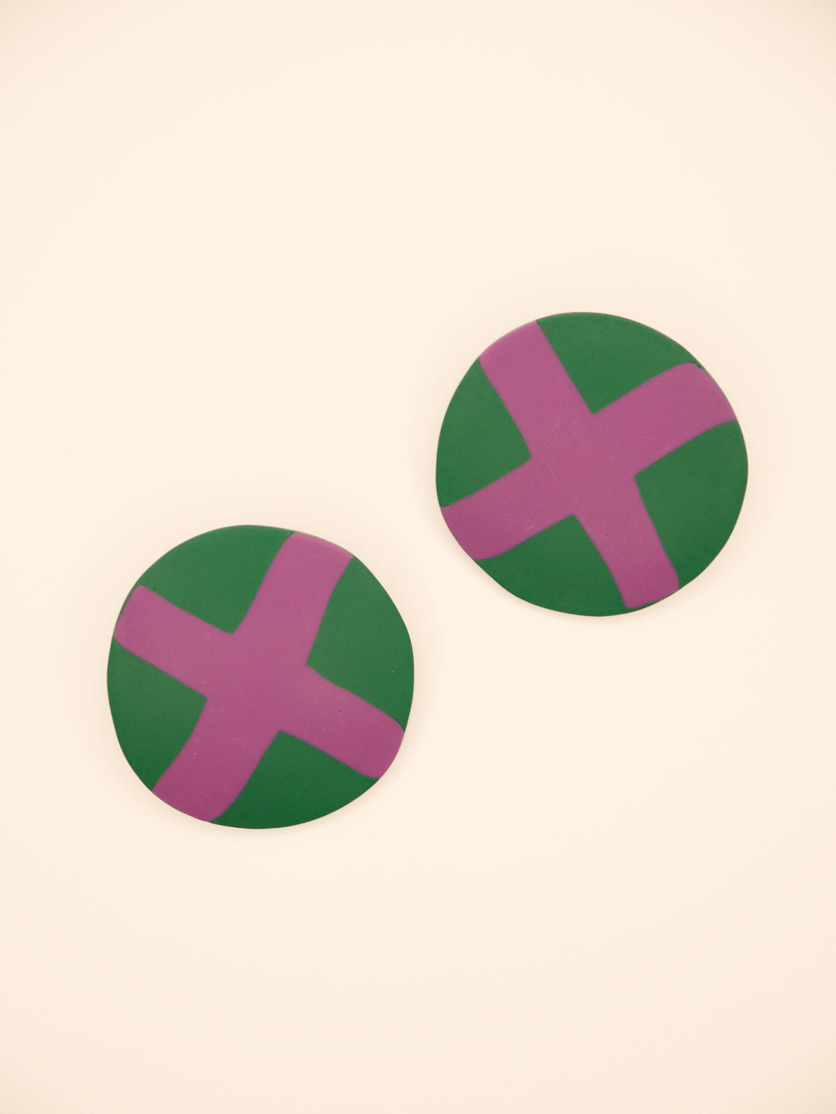 Two round green stud earrings with lilac 'X' design on a beige background