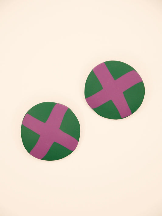 Two round green stud earrings with lilac 'X' design on a beige background