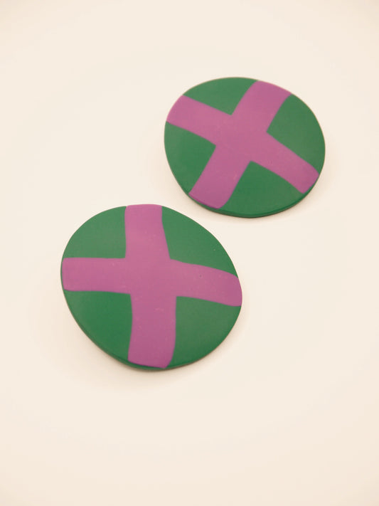 Two round green stud earrings with lilac 'X' design on a beige background