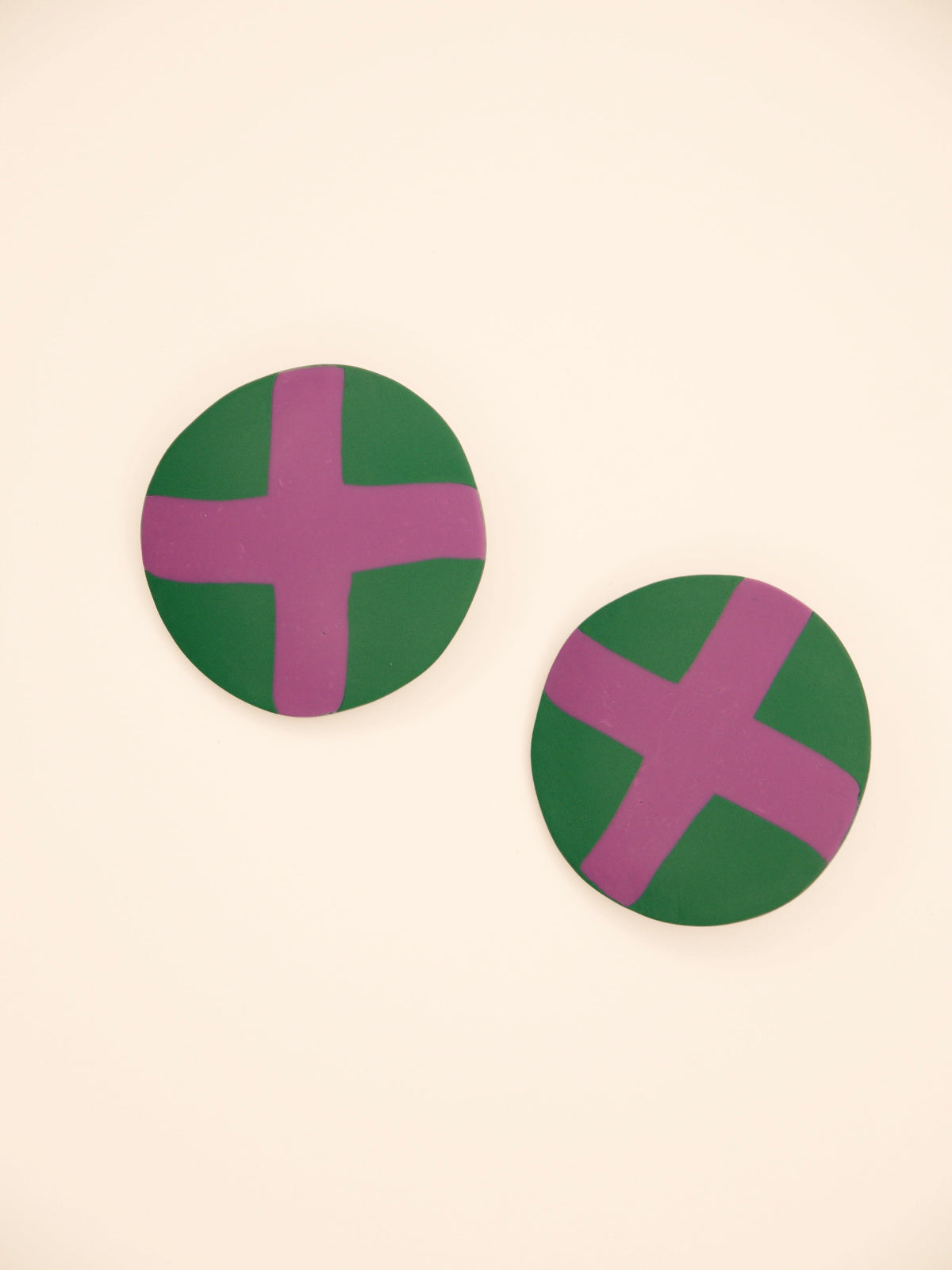 Two round green stud earrings with lilac 'X' design on a beige background