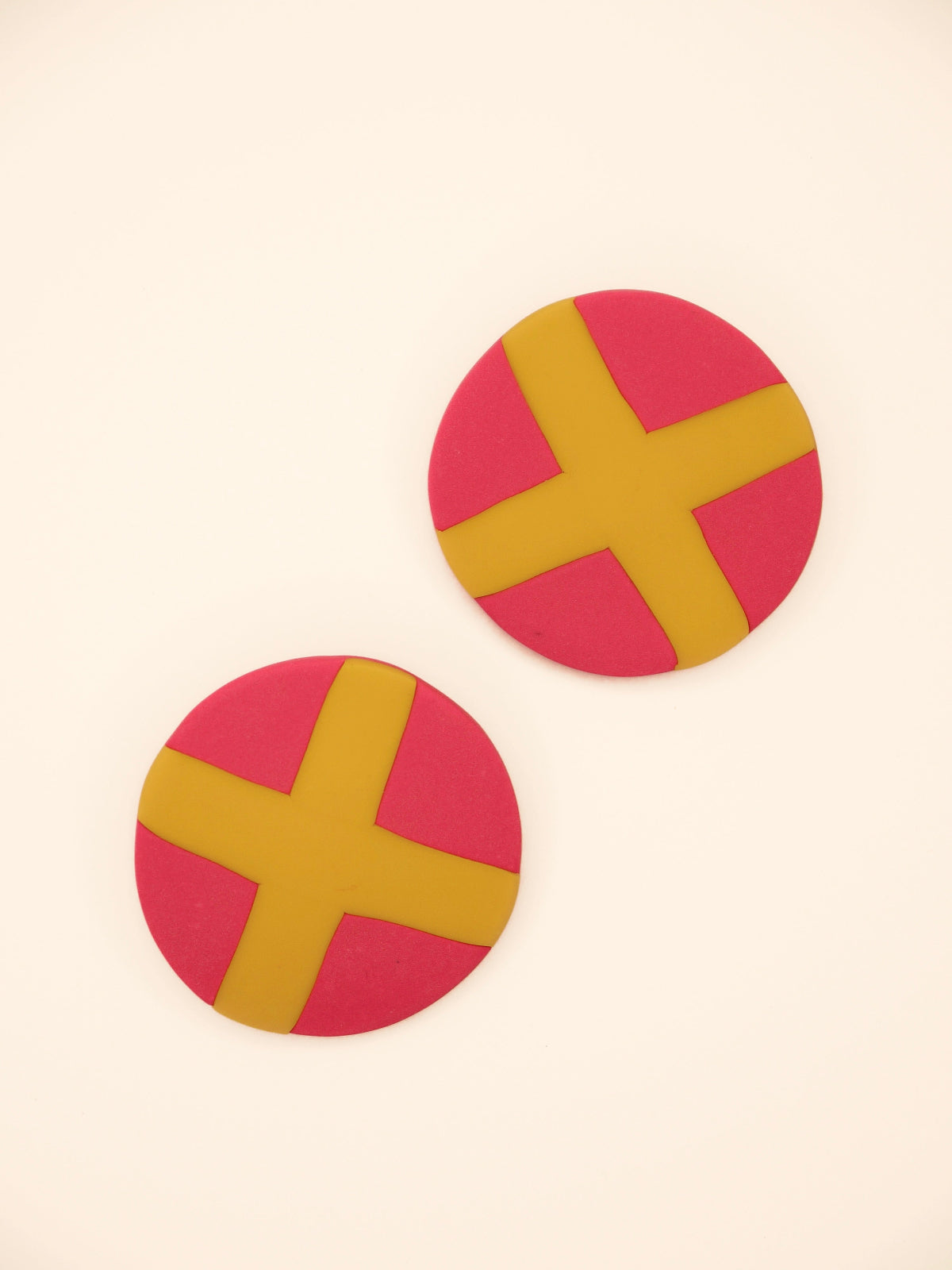Two round pink stud earrings with yellow 'X' design on a beige background