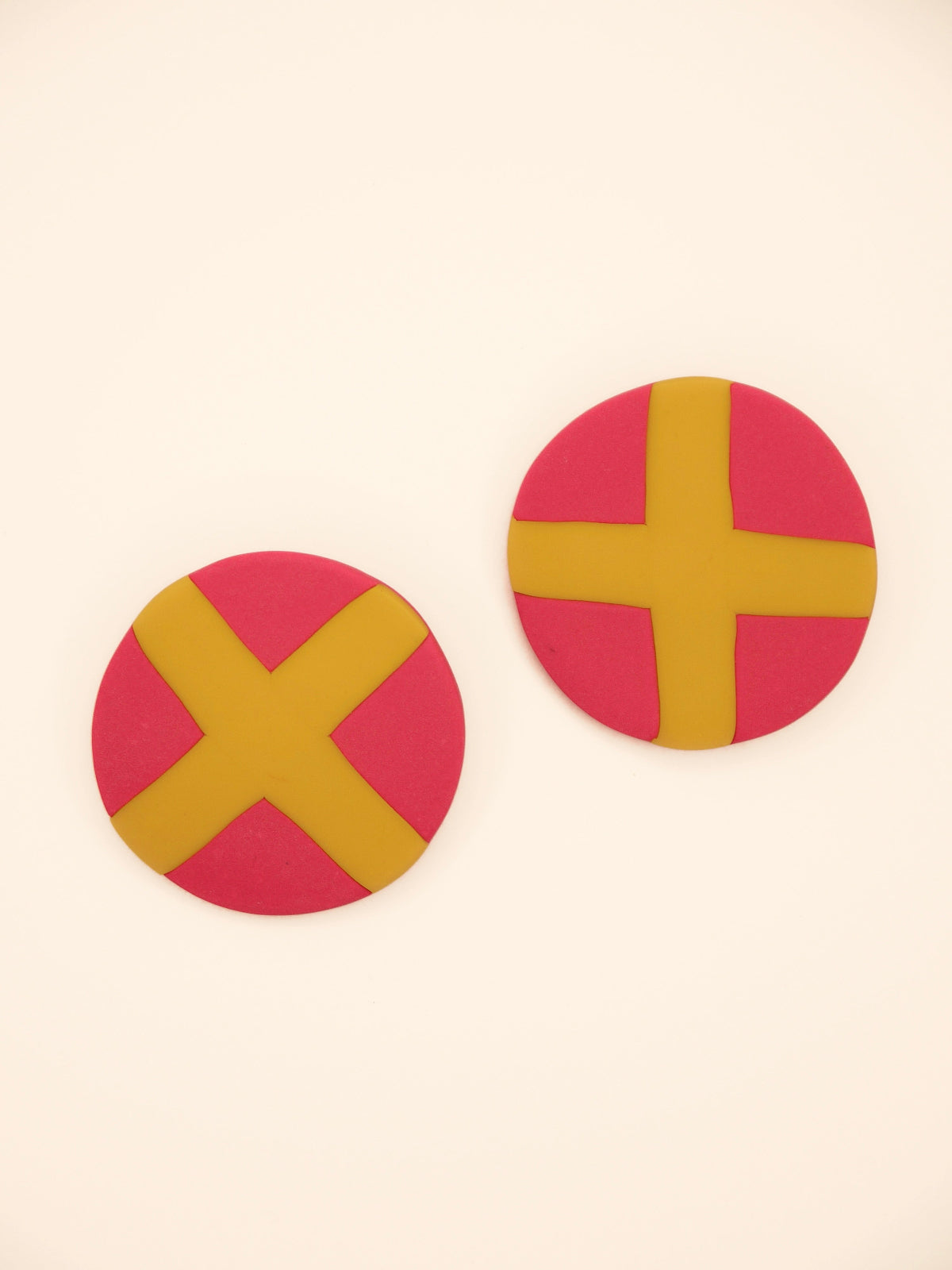 Two round pink stud earrings with yellow 'X' design on a beige background