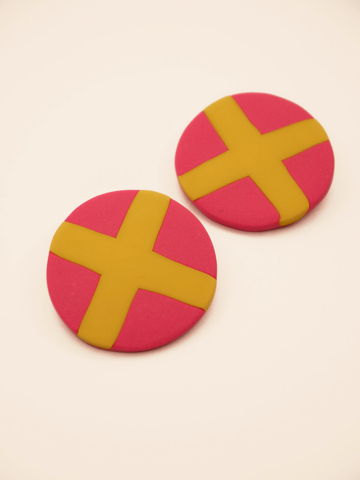 Two round pink stud earrings with yellow 'X' design on a beige background