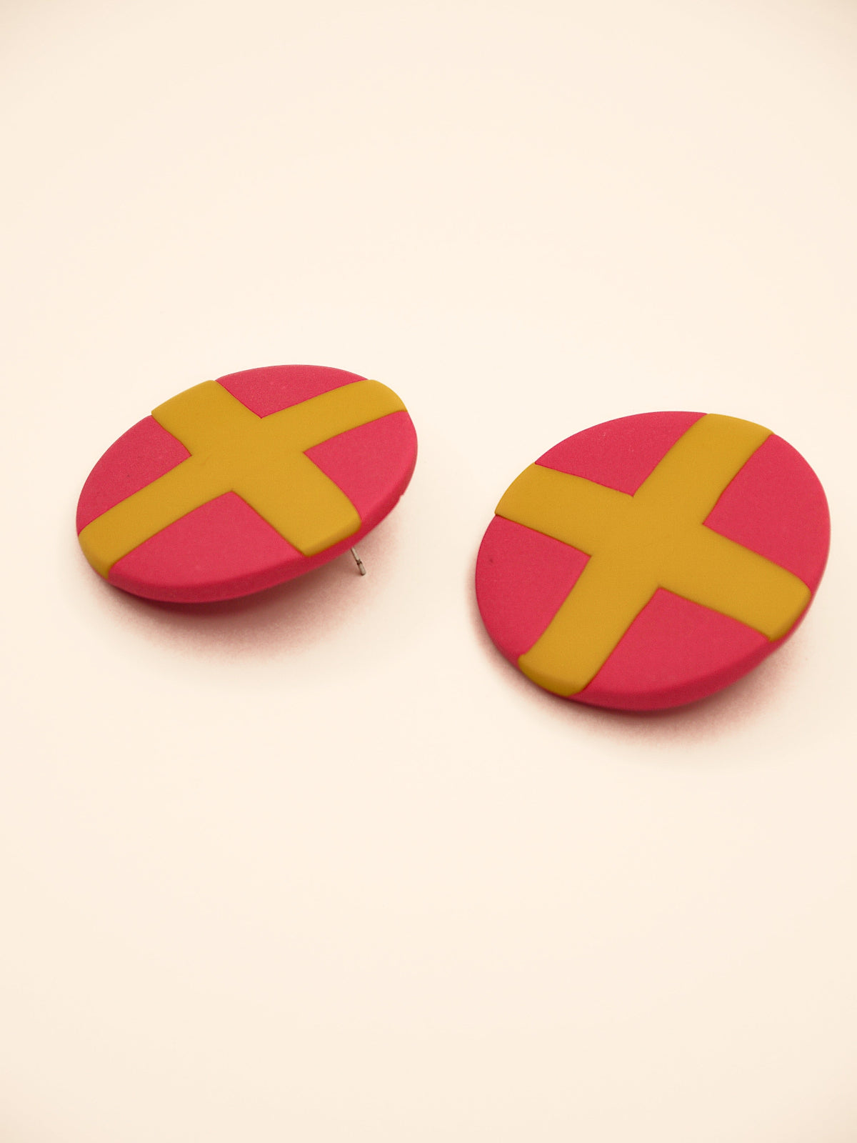 Two round pink stud earrings with yellow 'X' design on a beige background