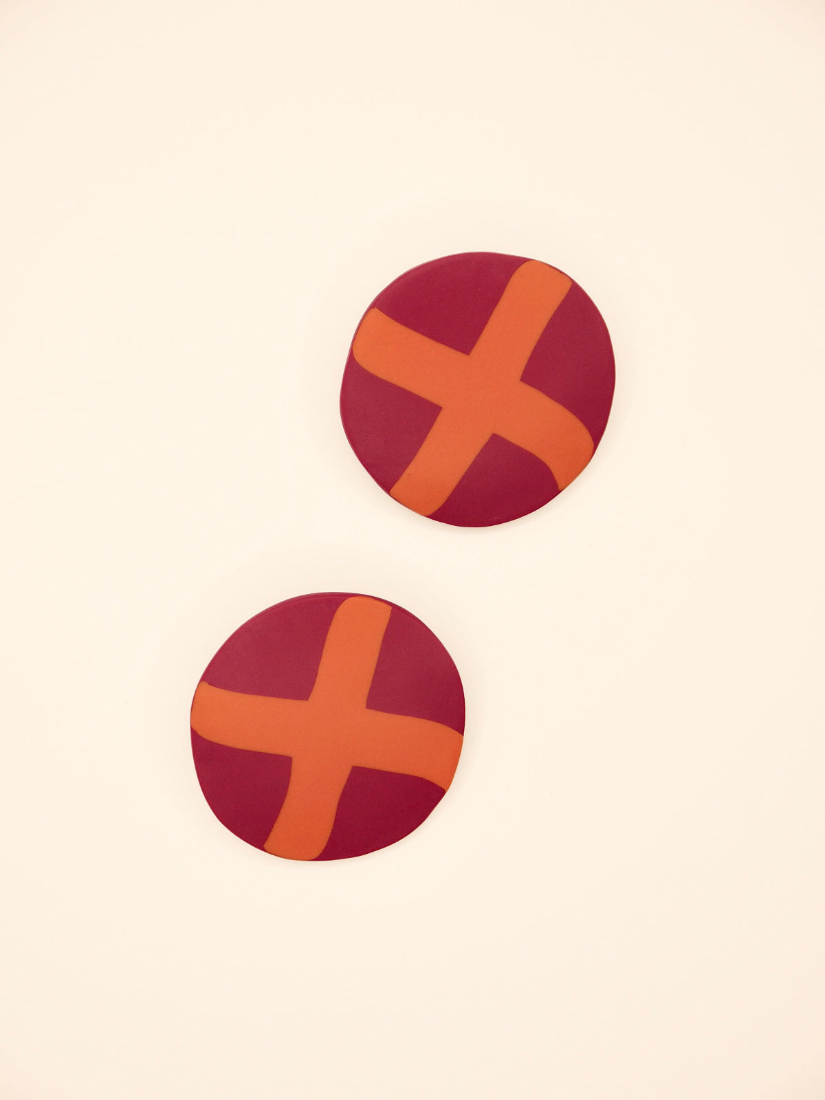 Two round purple stud earrings with orange 'X' design on a beige background
