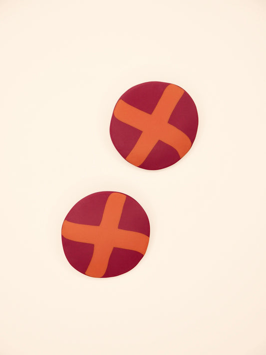 Two round purple stud earrings with orange 'X' design on a beige background