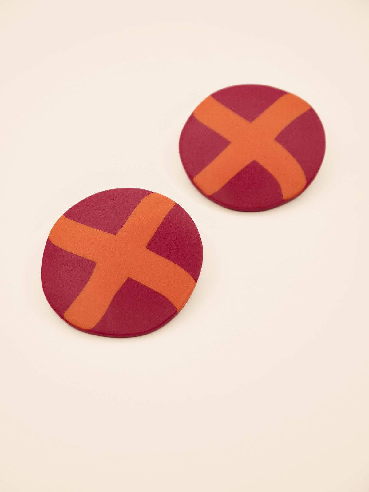 Two round purple stud earrings with orange 'X' design on a beige background
