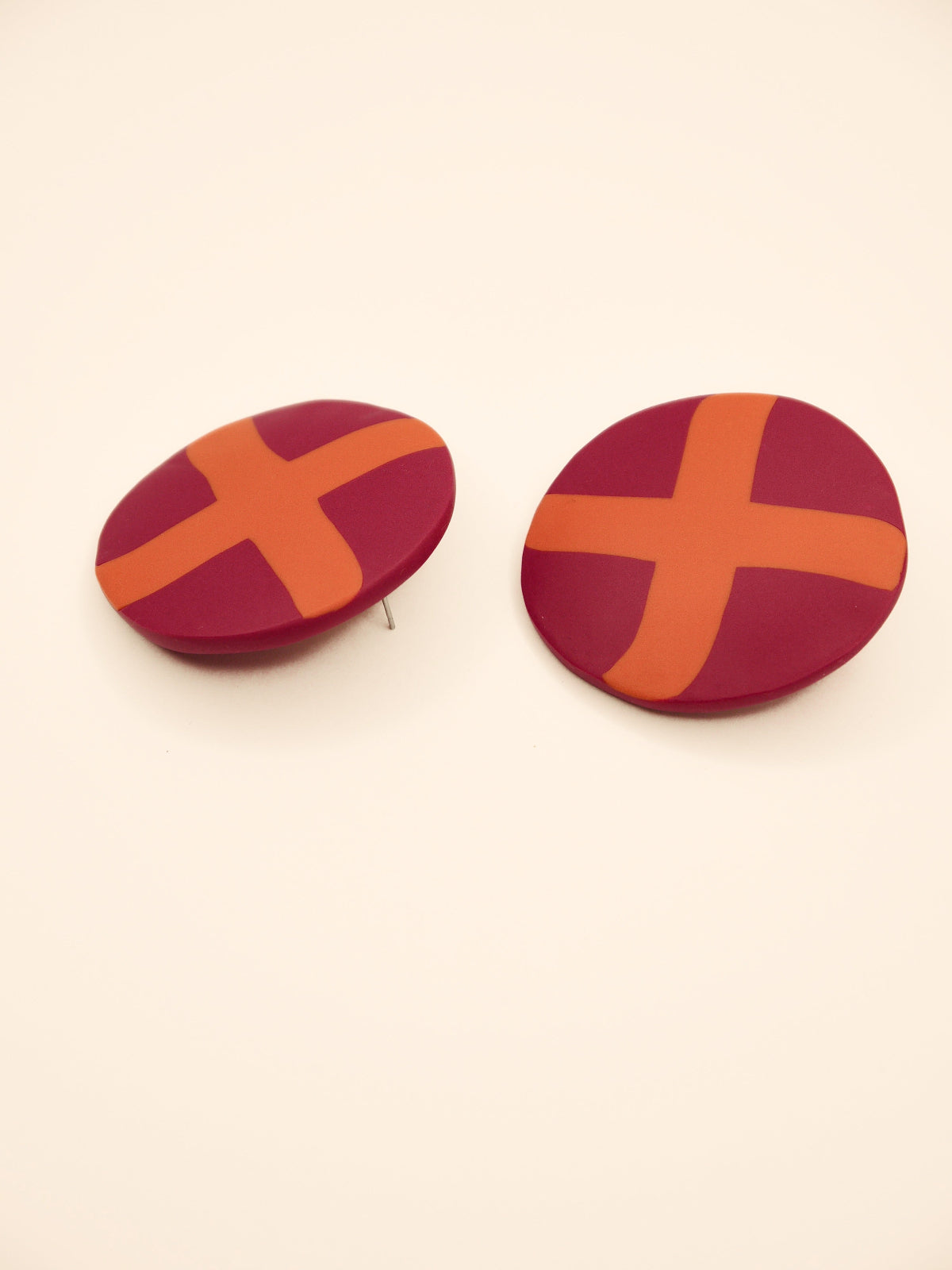 Two round purple stud earrings with orange 'X' design on a beige background