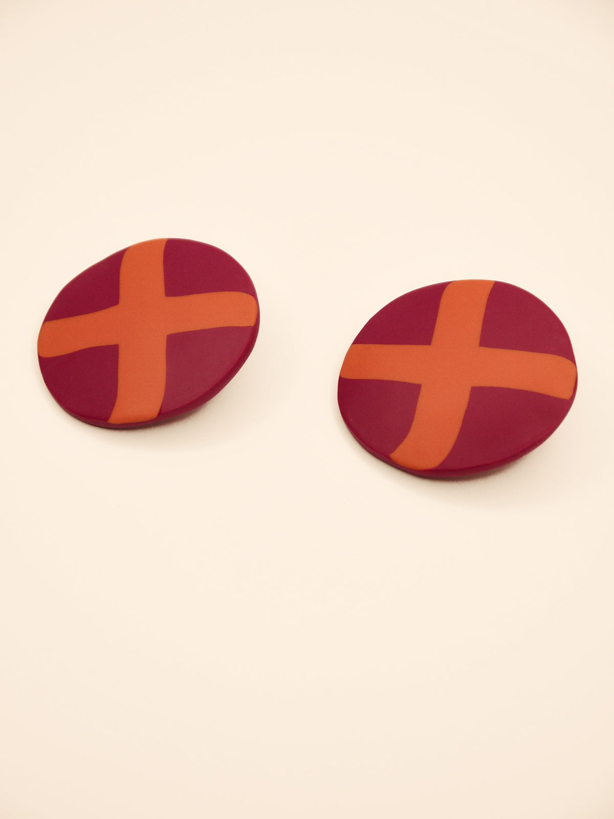 Two round purple stud earrings with orange 'X' design on a beige background