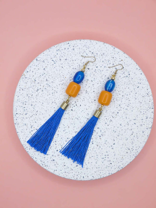 Blue and orange tassel earrings on a textured white platform with a pink background