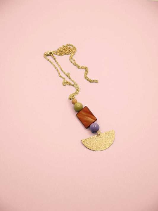 A bohemian-style pendant necklace featuring a gold chain, a textured gold semicircle pendant, a green bead, a purple bead, and a rectangular brown shell bead, displayed on a solid pink background.