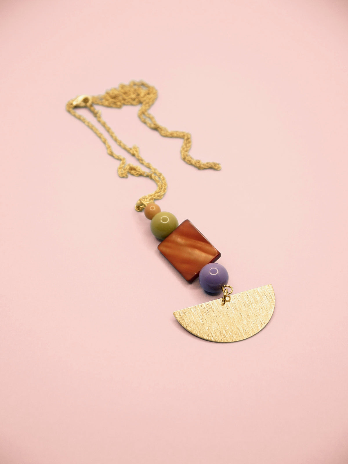 A bohemian-style geometric pendant necklace featuring a gold chain, a textured gold semicircle, a brown mother-of-pearl rectangle, a green oval bead, and a purple round bead, displayed on a soft pink background.