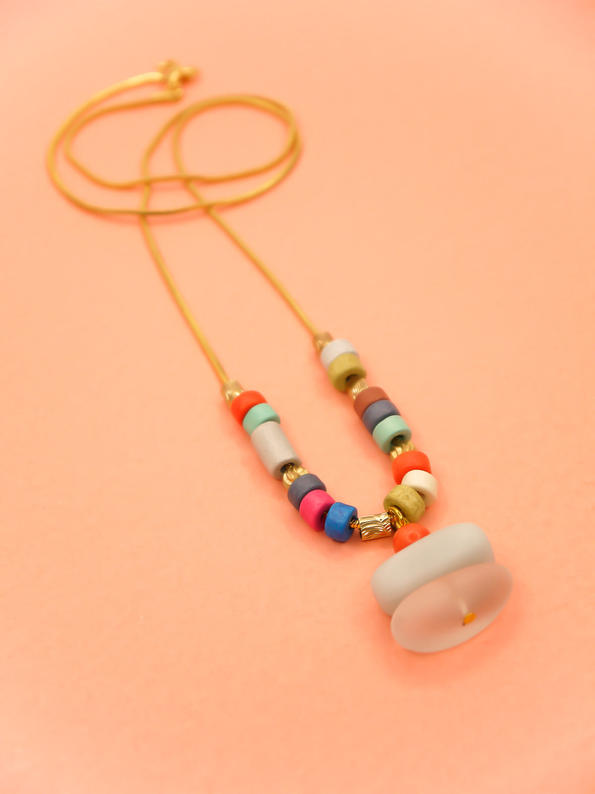 Crystal gemstone pendant necklace with a frosted crystal bead, stacked with an amazonite and coral bead, featuring multi coloured ceramic tube beads with textured brass tube beads hung on a gold plated snake chain