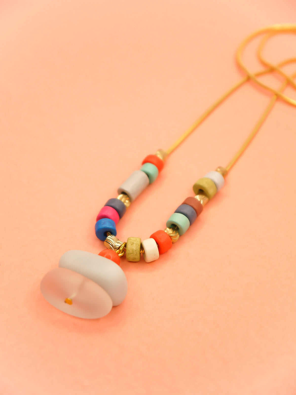 Crystal gemstone pendant necklace with a frosted crystal bead, stacked with an amazonite and coral bead, featuring multi coloured ceramic tube beads with textured brass tube beads hung on a gold plated snake chain