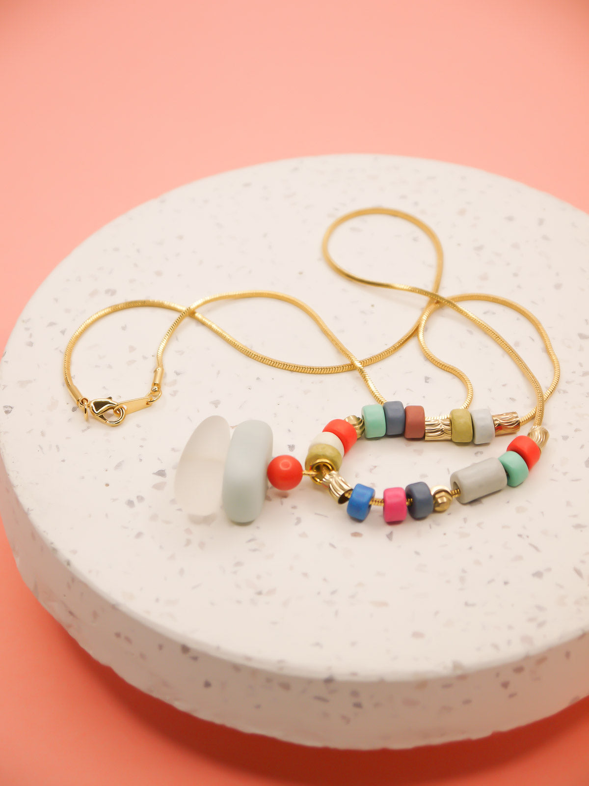 Crystal gemstone pendant necklace with a frosted crystal bead, stacked with an amazonite and coral bead, featuring multi coloured ceramic tube beads with textured brass tube beads hung on a gold plated snake chain