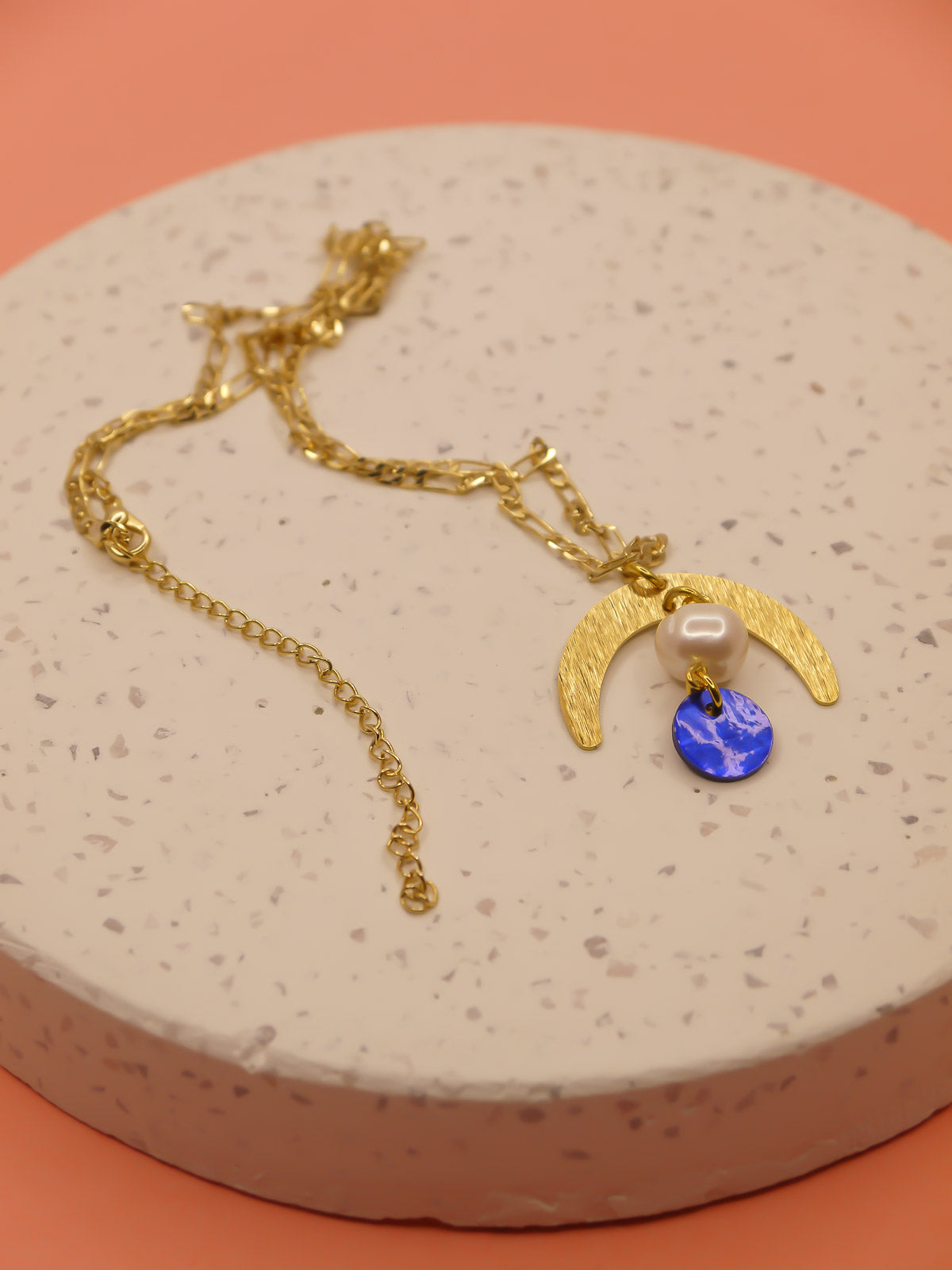 Geometric lightweight pendant necklace with a textured brass arch semi circle with a pearl and blue round shell disk hung from the arch on a faceted gold curb chain with extender.