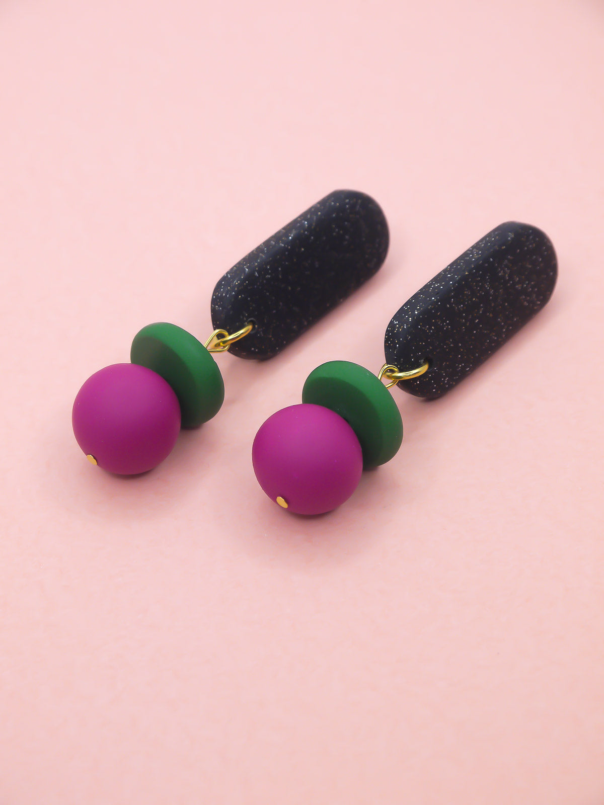 Colourful earrings with green, pink, and black components on a pink background