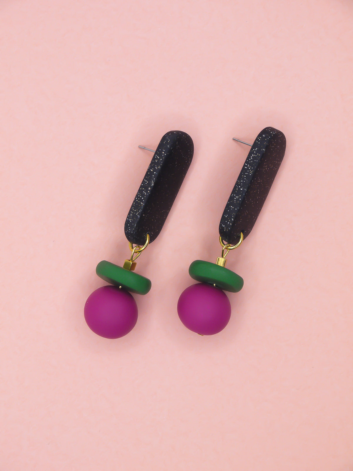 Colourful earrings with green, pink, and black components on a pink background