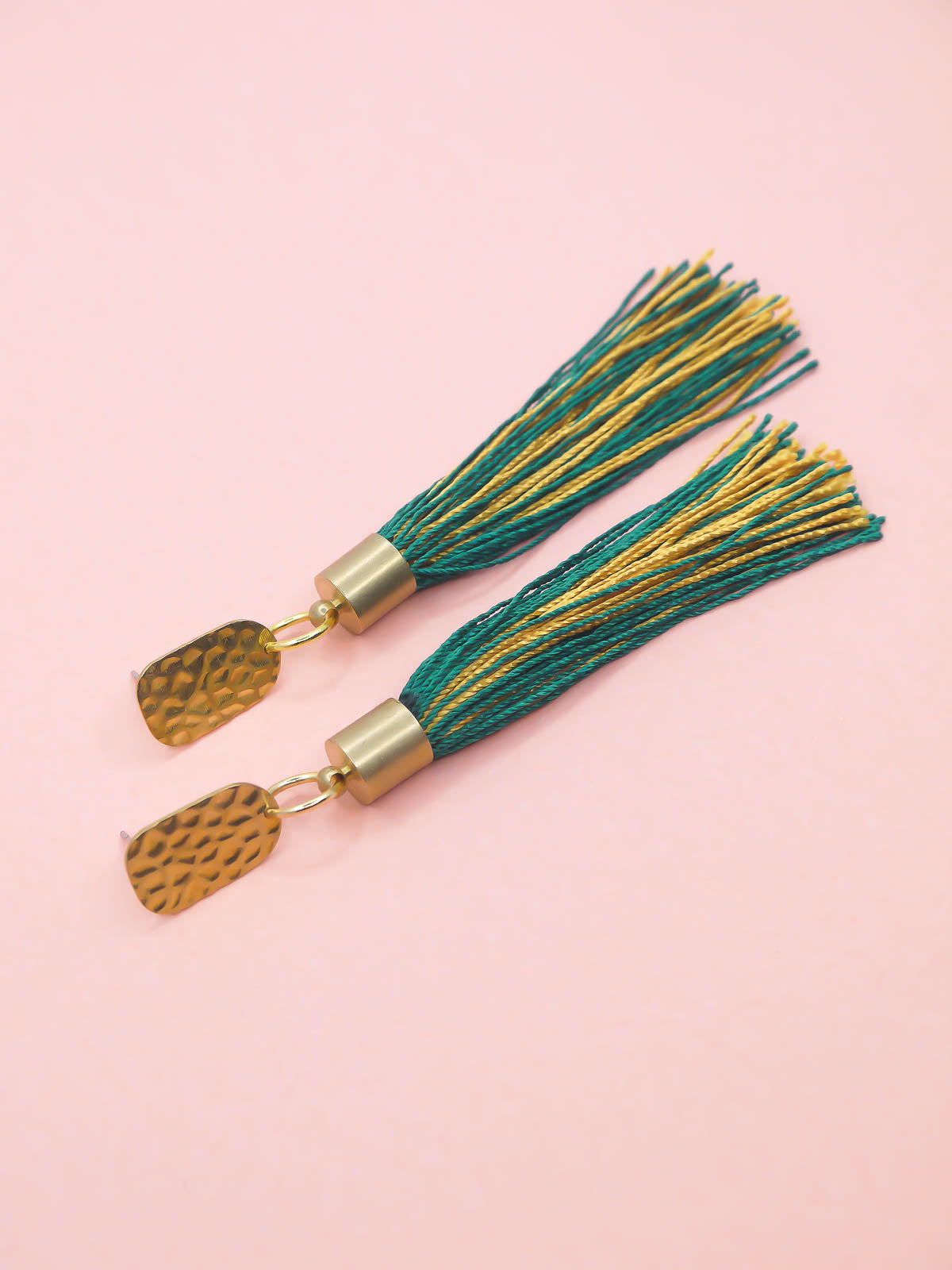 Tassel earrings with green and gold tassels and a textured brass stud on a pink background