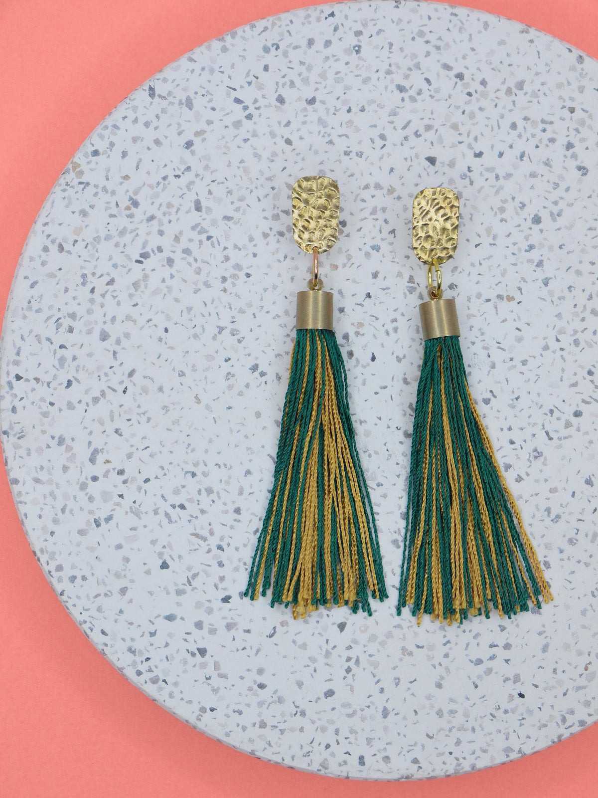 Green and gold tassel earrings with a textured brass stud on a marbled white surface with a pink background