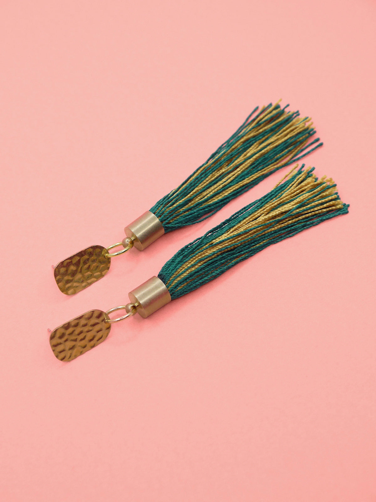 Tassel earrings with green and gold tassels and a textured brass stud on a pink background