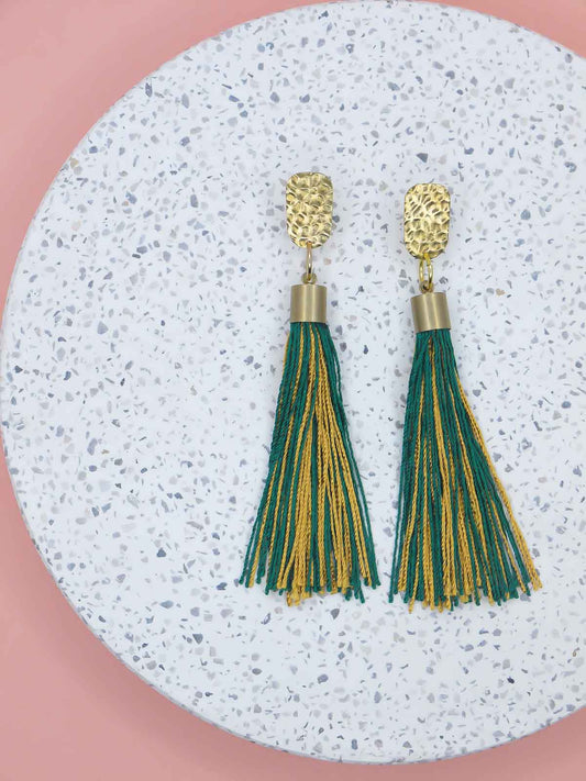 Green and gold tassel earrings with a textured brass stud on a marbled white surface with a pink background