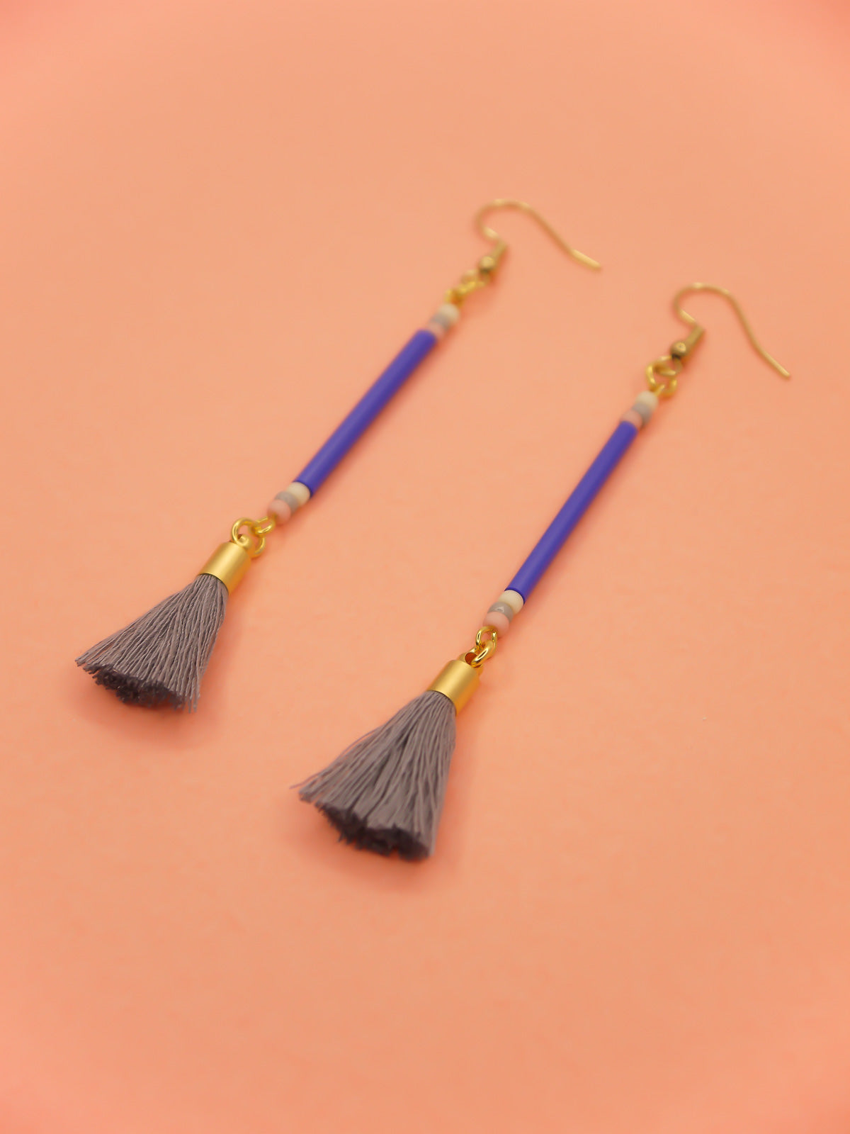 Long delicate bead stick dangle earrings featuring blue tube beads with contrasting seed beads in pink, white & grey, paired with a grey cotton tassel & hung from gold plated nickel free ear wires