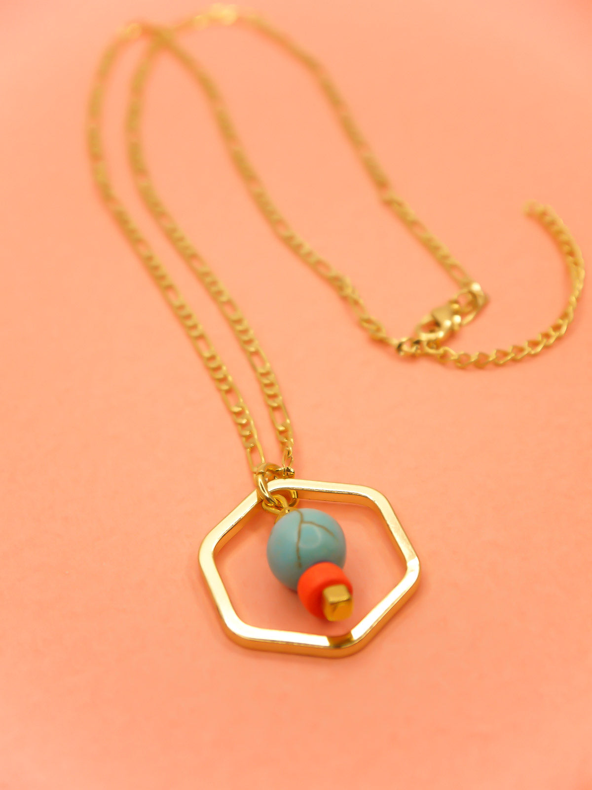 Geometric necklace comprising of a gold plated hexagon charm with a turquoise gemstone bead in the centre with an orange ceramic and brass square bead. Hung from a 24k shiny gold faceted curb chain with extender