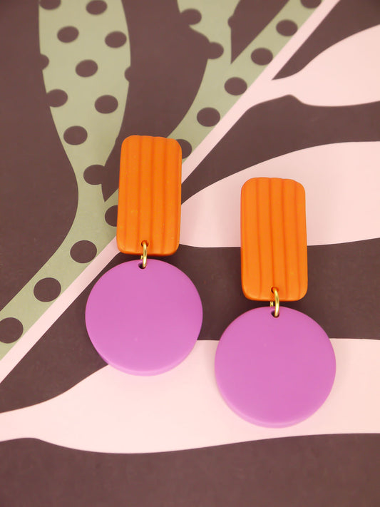 Pair of geometric earrings with orange striped rectangular tops and purple round bottoms on a patterned background.
