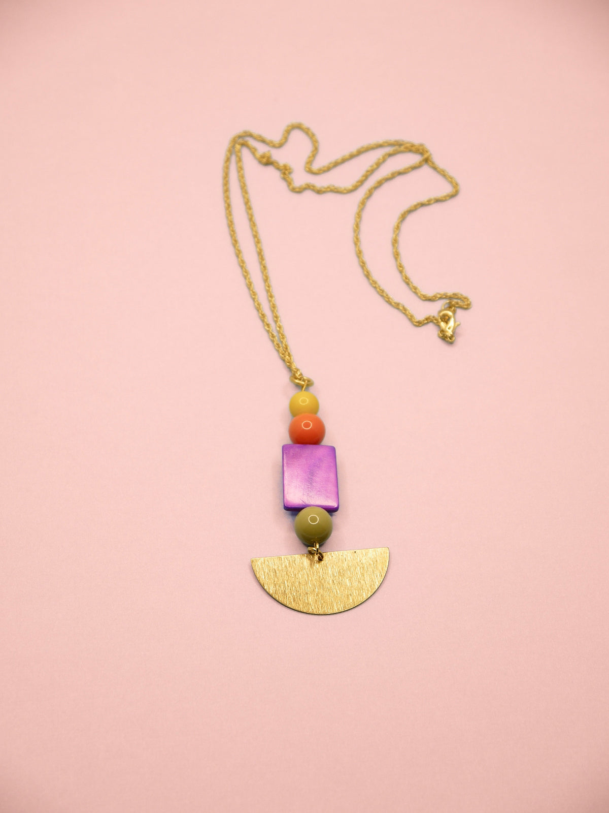A necklace featuring a gold chain, a gold-tone semicircle pendant, a purple square bead, a green bead, and small orange and yellow beads, displayed on a solid pink background.
