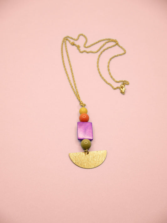 A necklace featuring a gold chain, a gold-tone semicircle pendant, a purple square bead, a green bead, and small orange and yellow beads, displayed on a solid pink background.