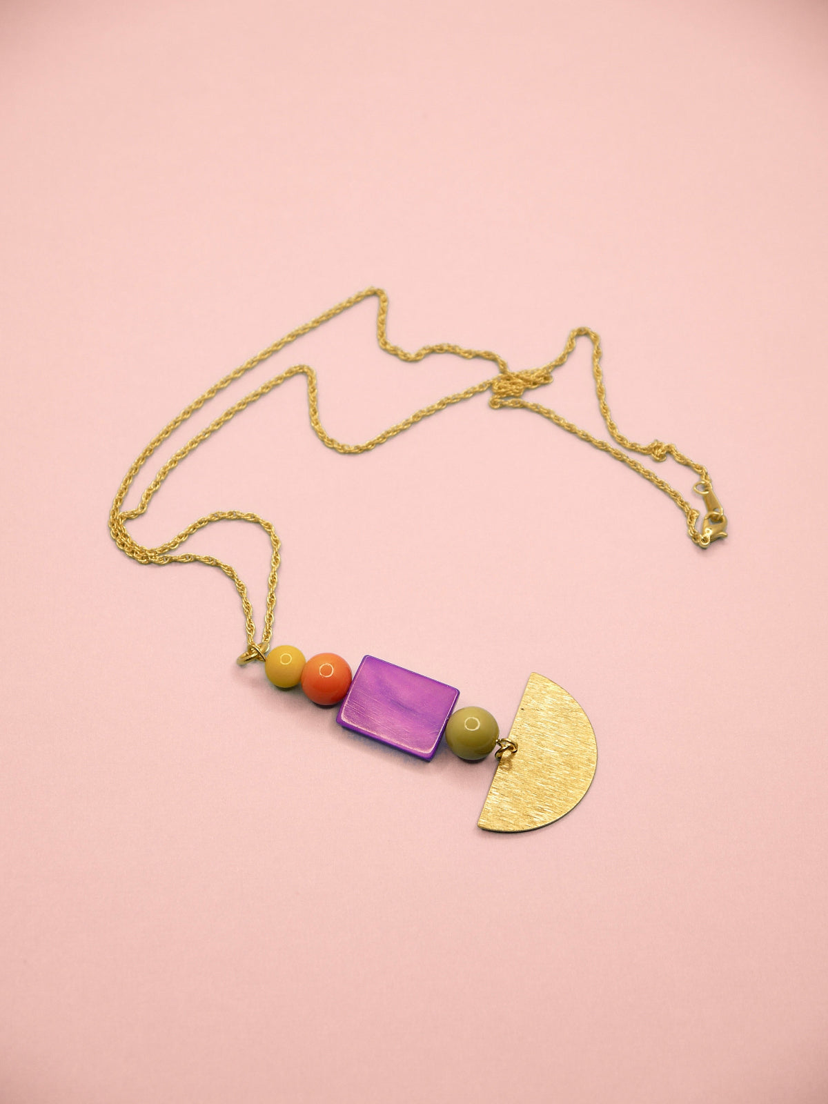 A gold chain necklace featuring a geometric pendant with a purple rectangular bead, three small colorful beads, a green oval bead, and a textured gold semicircle pendant, displayed on a solid pink background.
