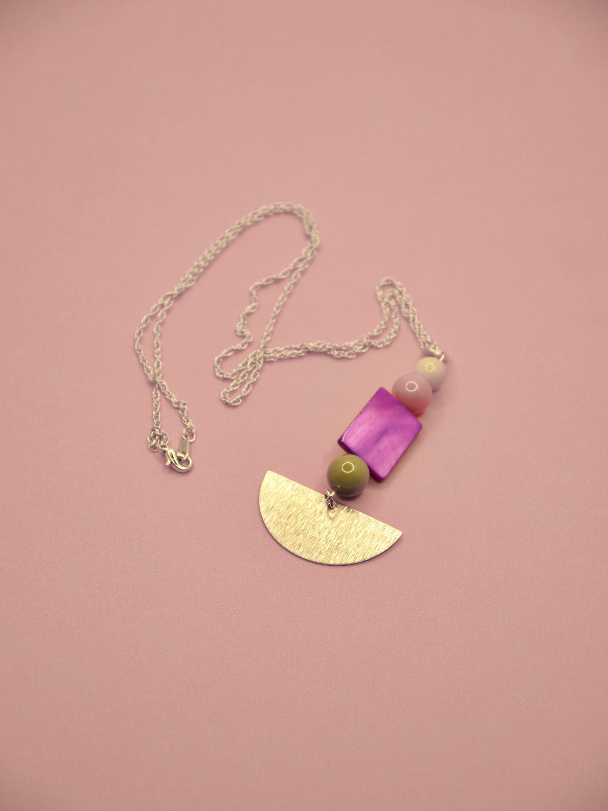 A bohemian-style pendant necklace featuring a textured gold semicircle, a pink and white beaded strand, and a green bead on a gold chain, displayed on a solid dusty pink background.
