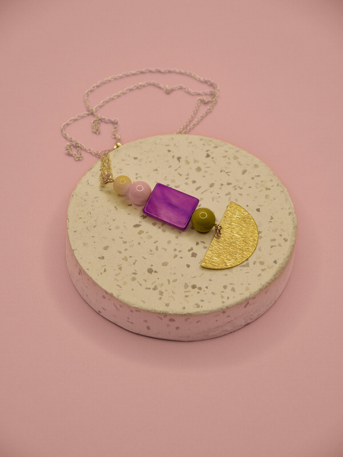 A handmade necklace with a gold chain, featuring a purple rectangular stone, a gold crescent moon, a green bead, and pastel pink and yellow beads, displayed on a round speckled pedestal against a solid pink background.