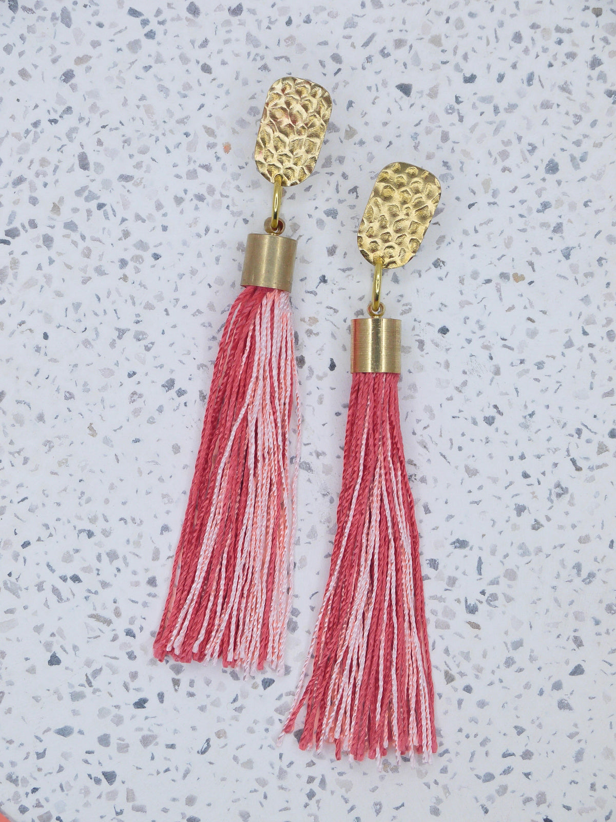 Red & pink tassel dangle earrings with a textured brass stud on a marble background