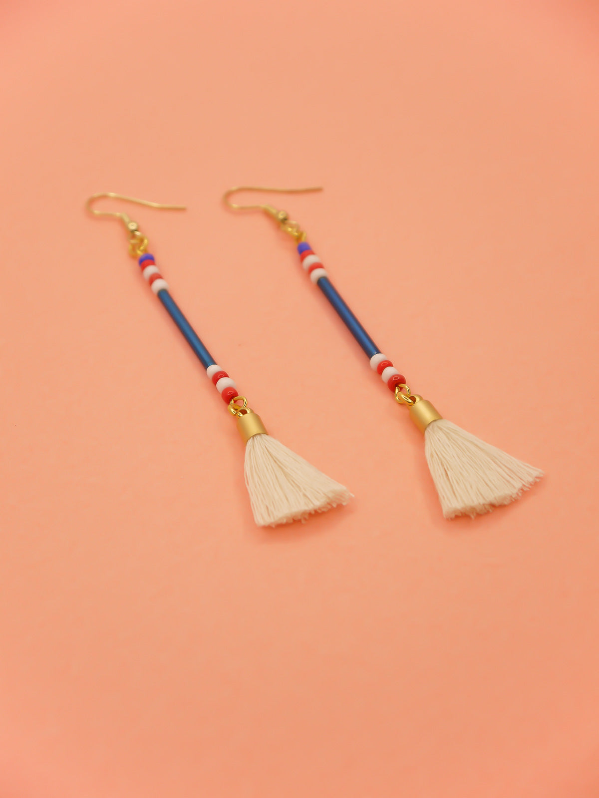 Lightweight delicate long beaded dangle earrings with a blue metallic tube bead and contrasting seed beads in red, white & blue, paired with a white cotton tassel. Hung from gold plated nickel free ear wires. 