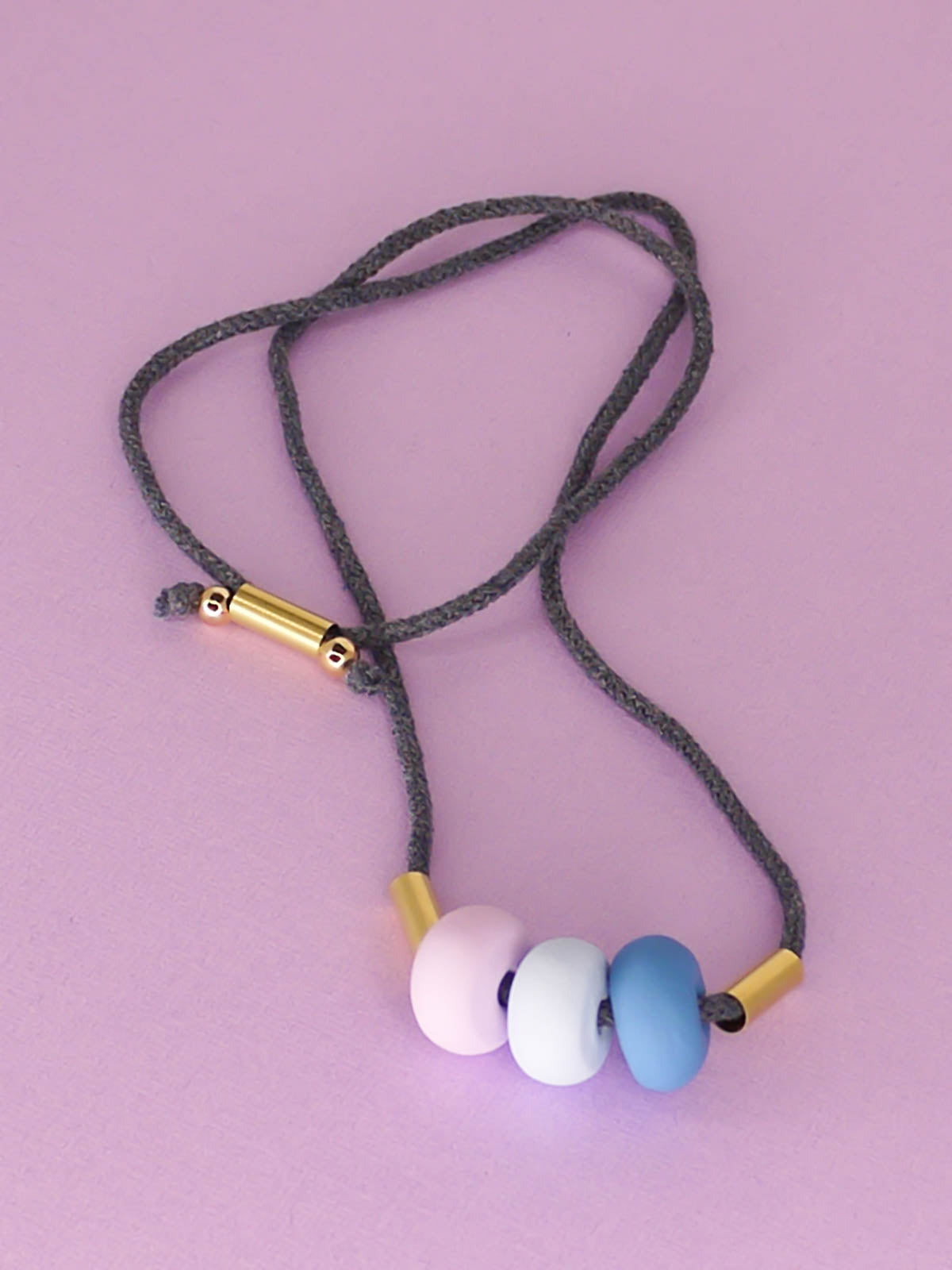 Lightweight statement bead necklace made up of grey cotton rope which is adjustable through a brass slider so it can be worn long or short. Comprised of 3 handmade polymer clay beads in blue, ice blue & lilac with 2 brass tube beads.