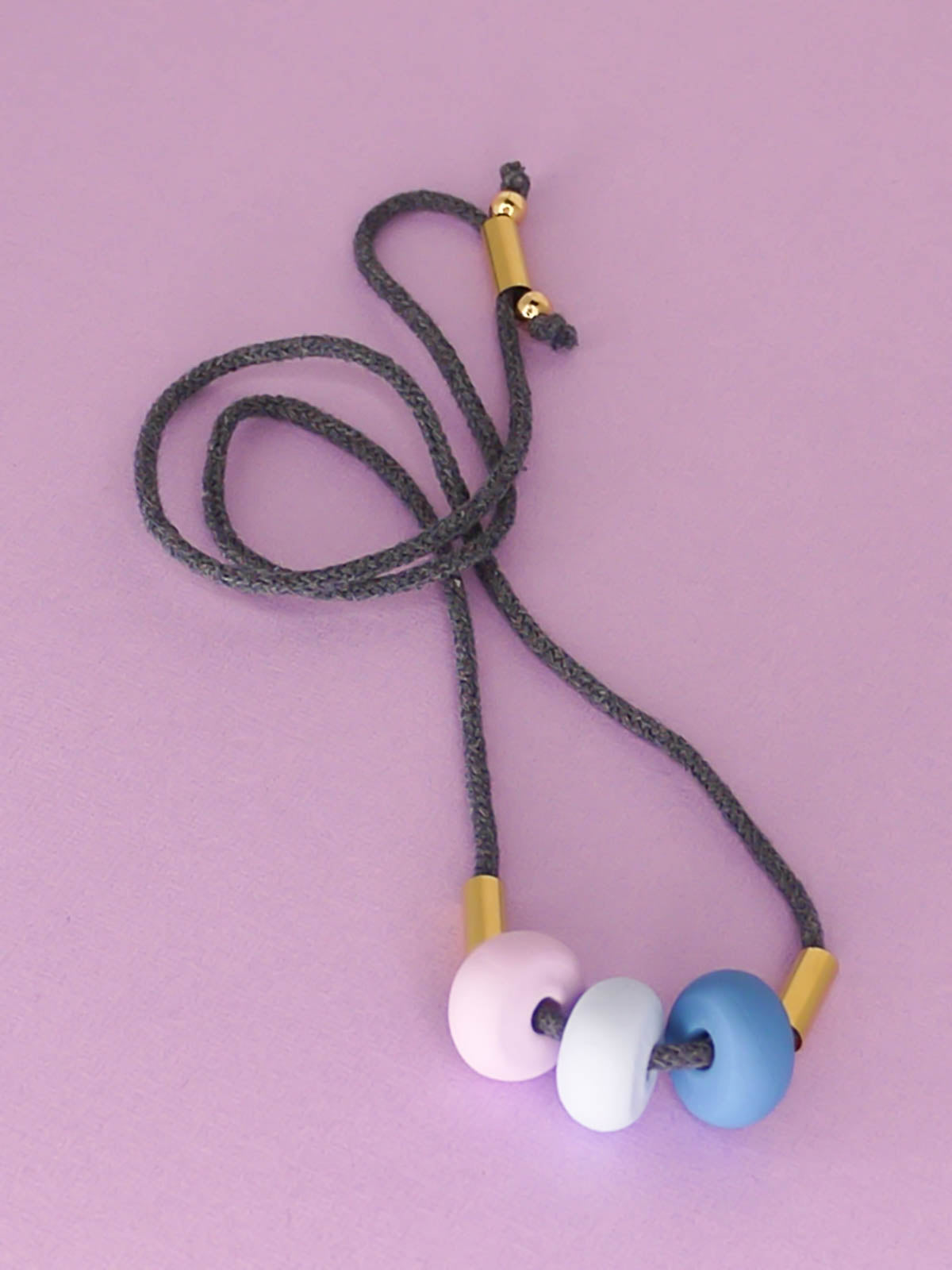 Lightweight statement bead necklace made up of grey cotton rope which is adjustable through a brass slider so it can be worn long or short. Comprised of 3 handmade polymer clay beads in blue, ice blue & lilac with 2 brass tube beads.