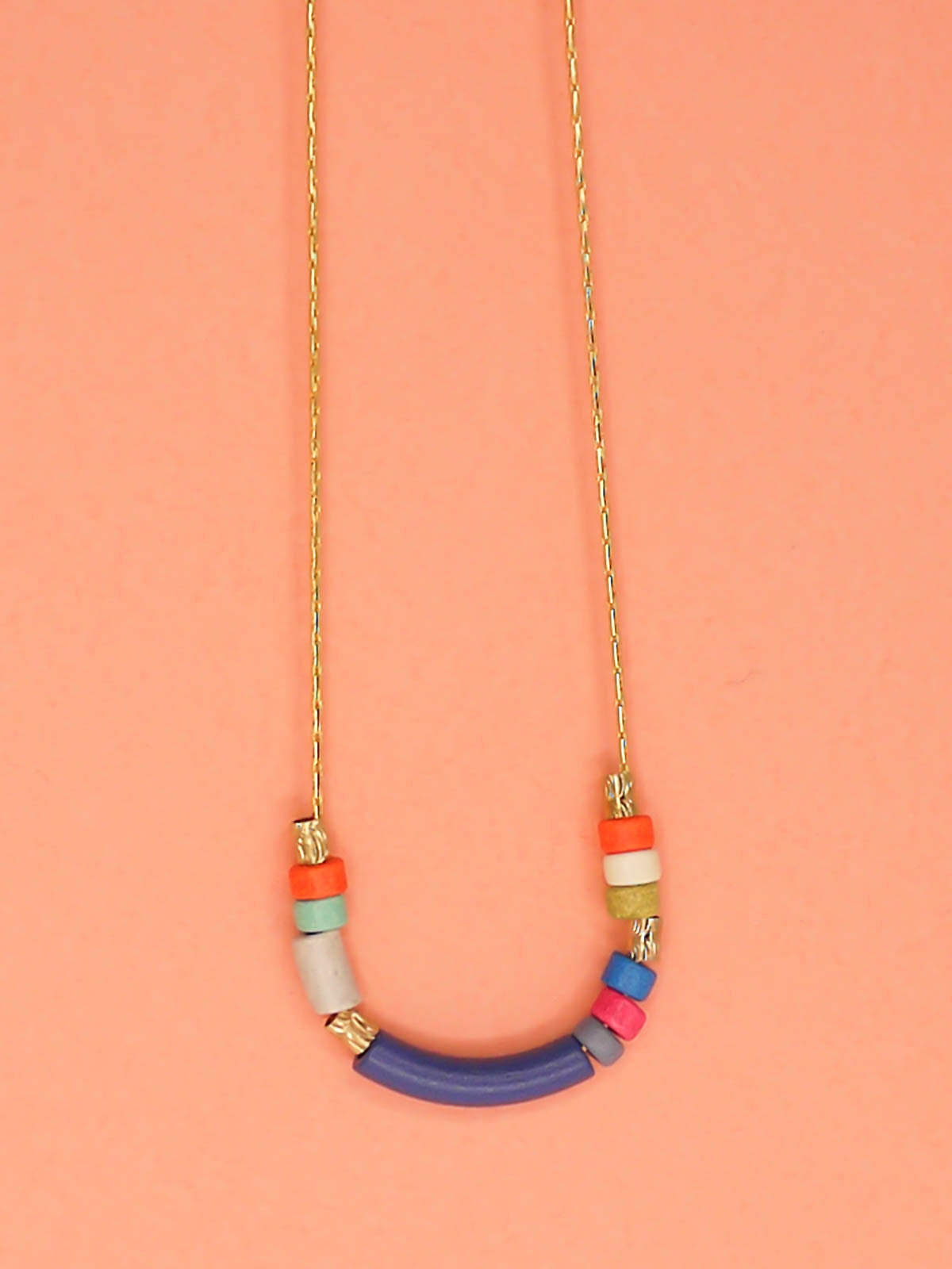 Delicate ceramic bead necklace with a blue curved bar bead, multicoloured ceramic tube beads and brass tube beads strung on a gold plated snake chain