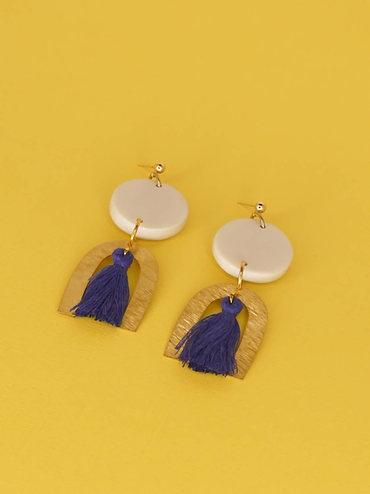 Geometric statement earrings with a textured brass arch and blue cotton tassel hung from a white polymer clay circle disk with gold plated ball studs