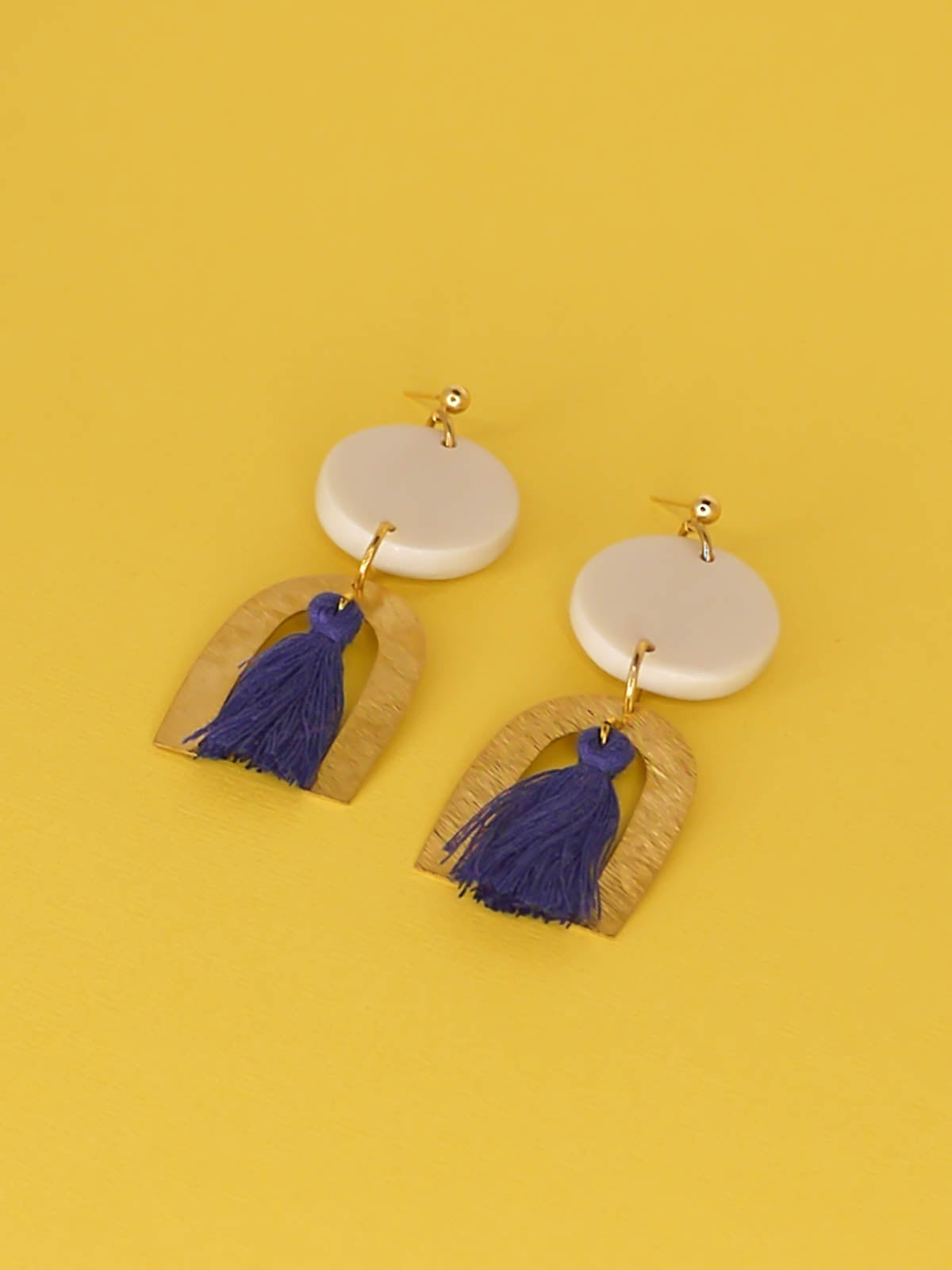 Geometric statement earrings with a textured brass arch and blue cotton tassel hung from a white polymer clay circle disk with gold plated ball studs