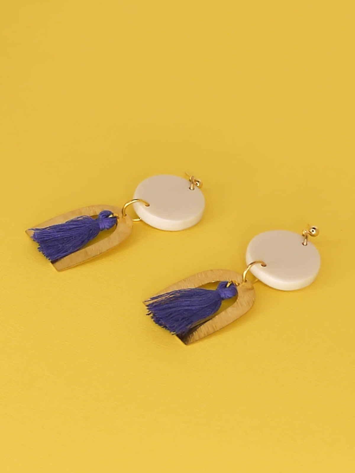 Geometric statement earrings with a textured brass arch and blue cotton tassel hung from a white polymer clay circle disk with gold plated ball studs