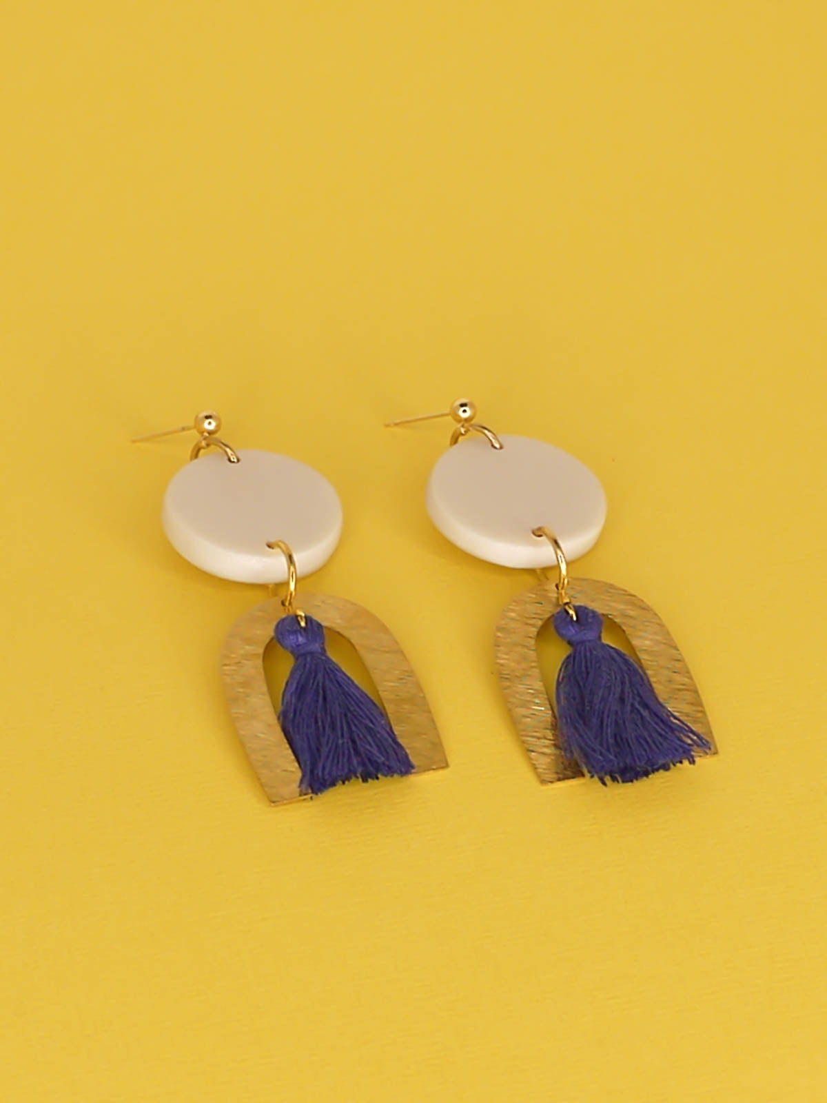 Geometric statement earrings with a textured brass arch and blue cotton tassel hung from a white polymer clay circle disk with gold plated ball studs